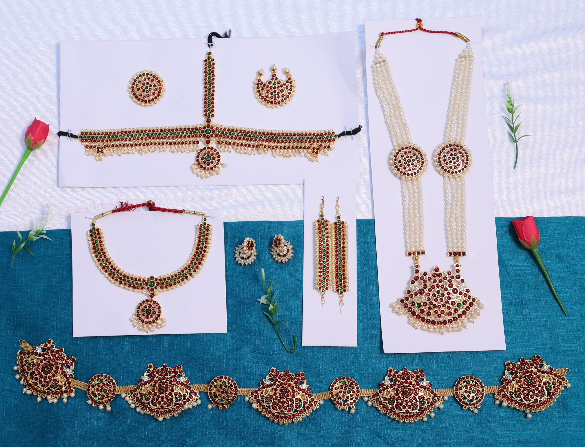 Wholesale Bharatanatyam temple jewellery set with Kemp red and green stones, including long pearl haram, choker, earrings, mattal, headpiece, and waist belt displayed on blue and white background