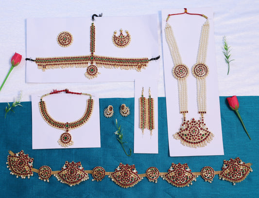Wholesale Bharatanatyam temple jewellery set with Kemp red and green stones, including long pearl haram, choker, earrings, mattal, headpiece, and waist belt displayed on blue and white background